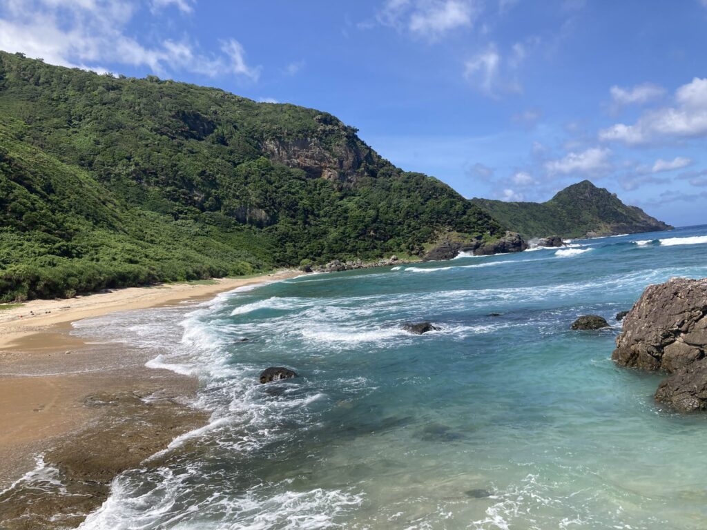 与路島の高原海岸。緑の山に囲まれた誰もいない砂浜に、透き通った青い波が打ち寄せる