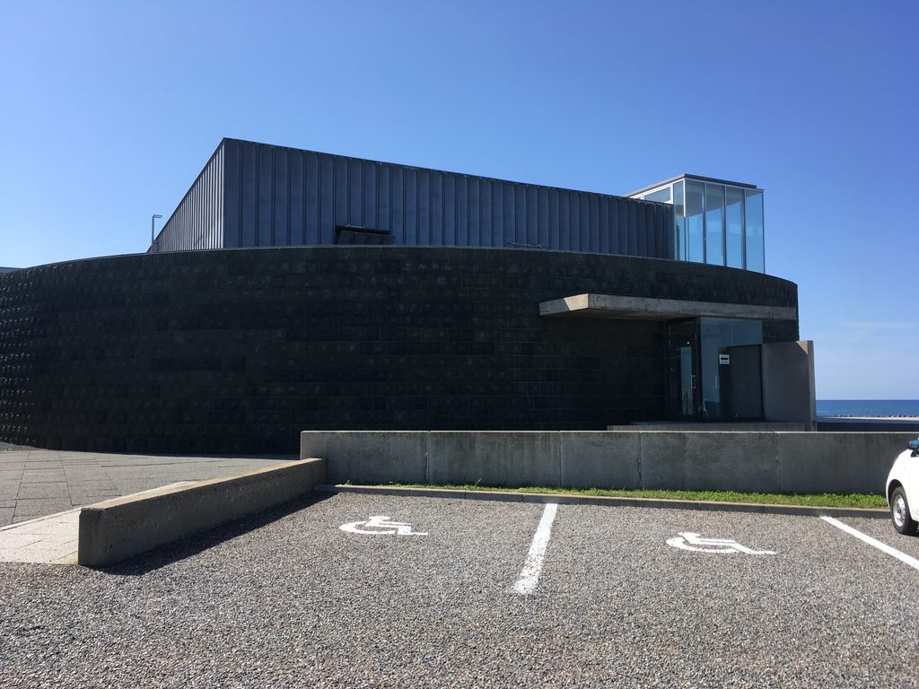 奥尻島津波館の外観|黒い壁の建物と駐車場、奥に海|奥尻島青苗