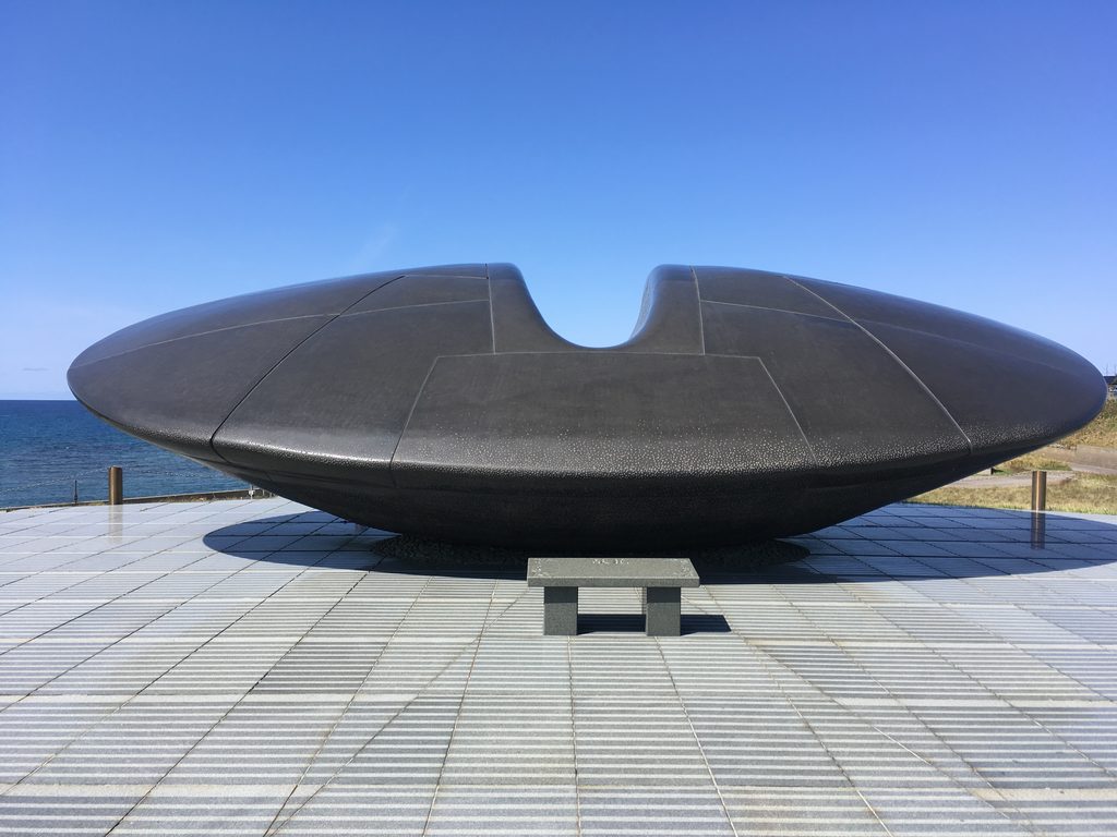 慰霊碑「時空翔」|黒御影石のくぼみをもつ碑が、海に面した石畳の広場に置かれている|奥尻島